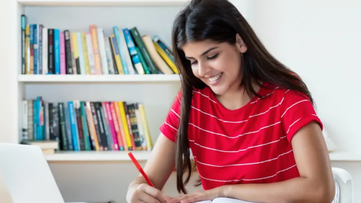 girl student in red shirt at desk, smiling, using laptop and writing