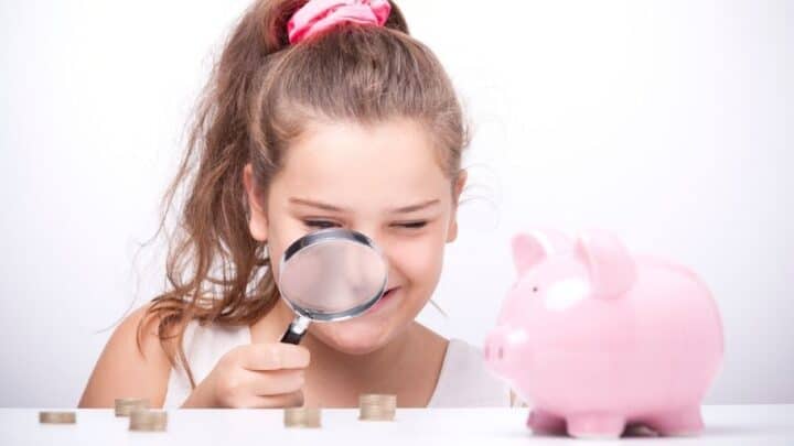 tween girl with magnifying glass looking at money and piggy bank