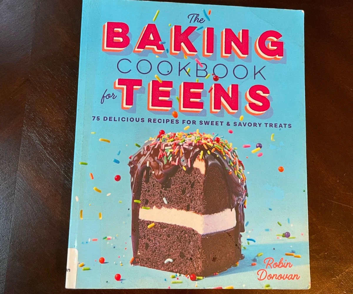 The Baking Cookbook for Teens blue color with giant chocolate piece of cake on it, on dark table