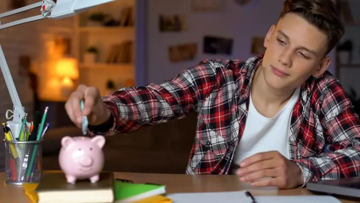 teen boy at desk putting money into piggy bank