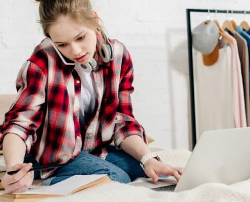 young teen working on a business plan for students worksheet on her bed, with laptop, books, and while on phone