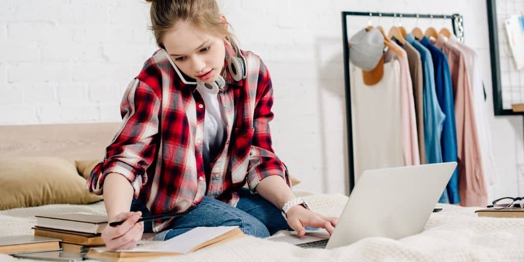 young teen working on a business plan for students worksheet on her bed, with laptop, books, and while on phone