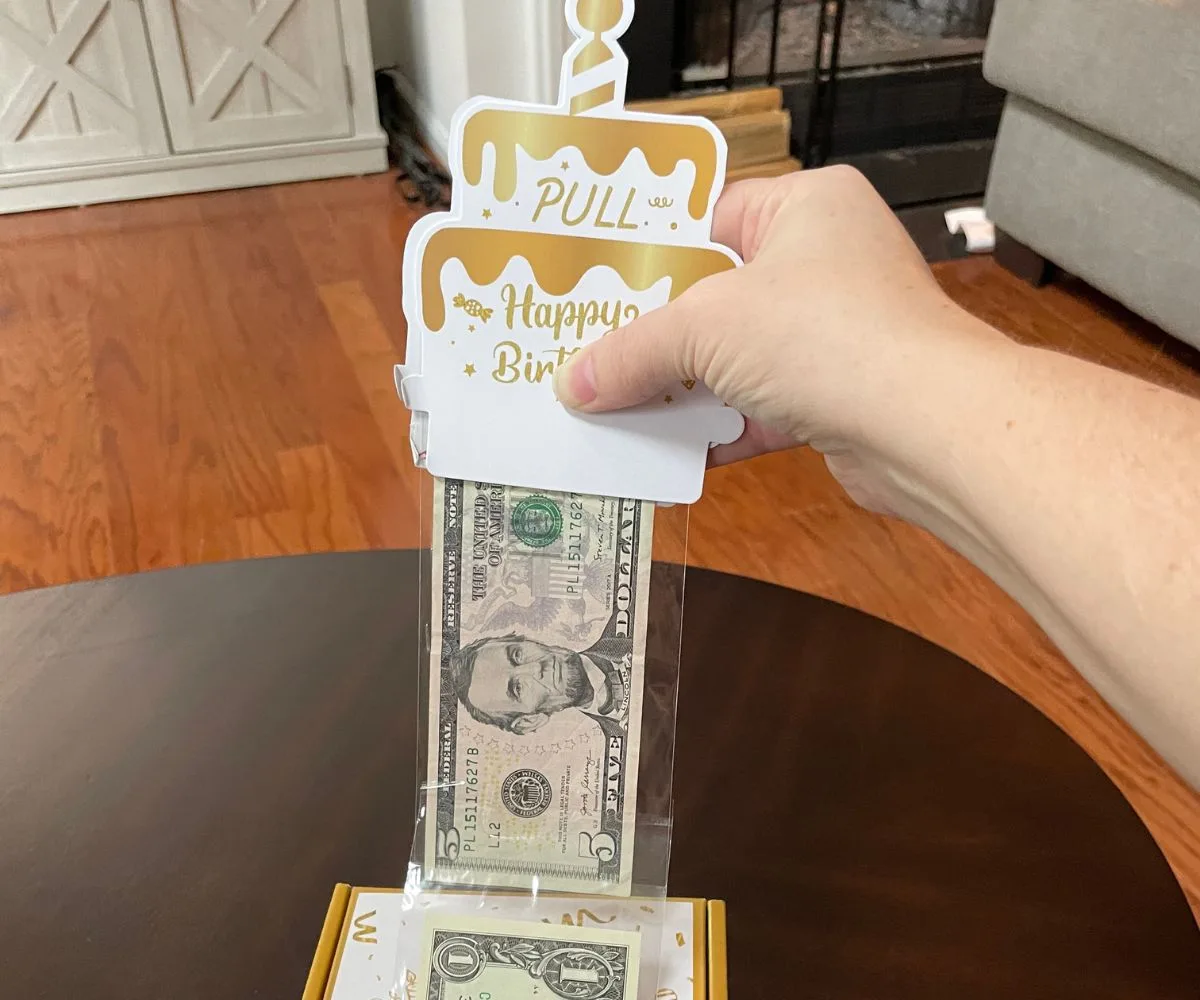 hand pulling gold and white birthday cake card up with money attached to it coming out of box