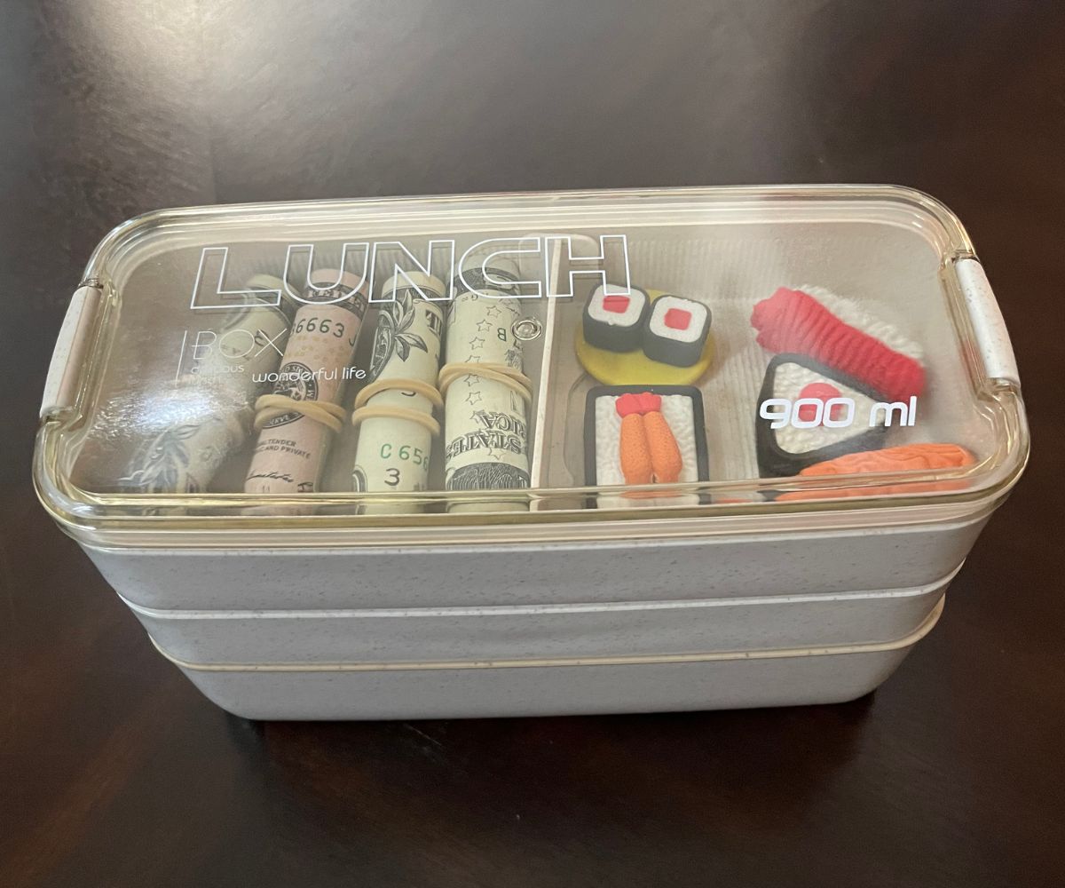 three-tier bento box khaki colored with rolled dollar and 5 dollar bills on one part of top and eraser sushi in other, closed