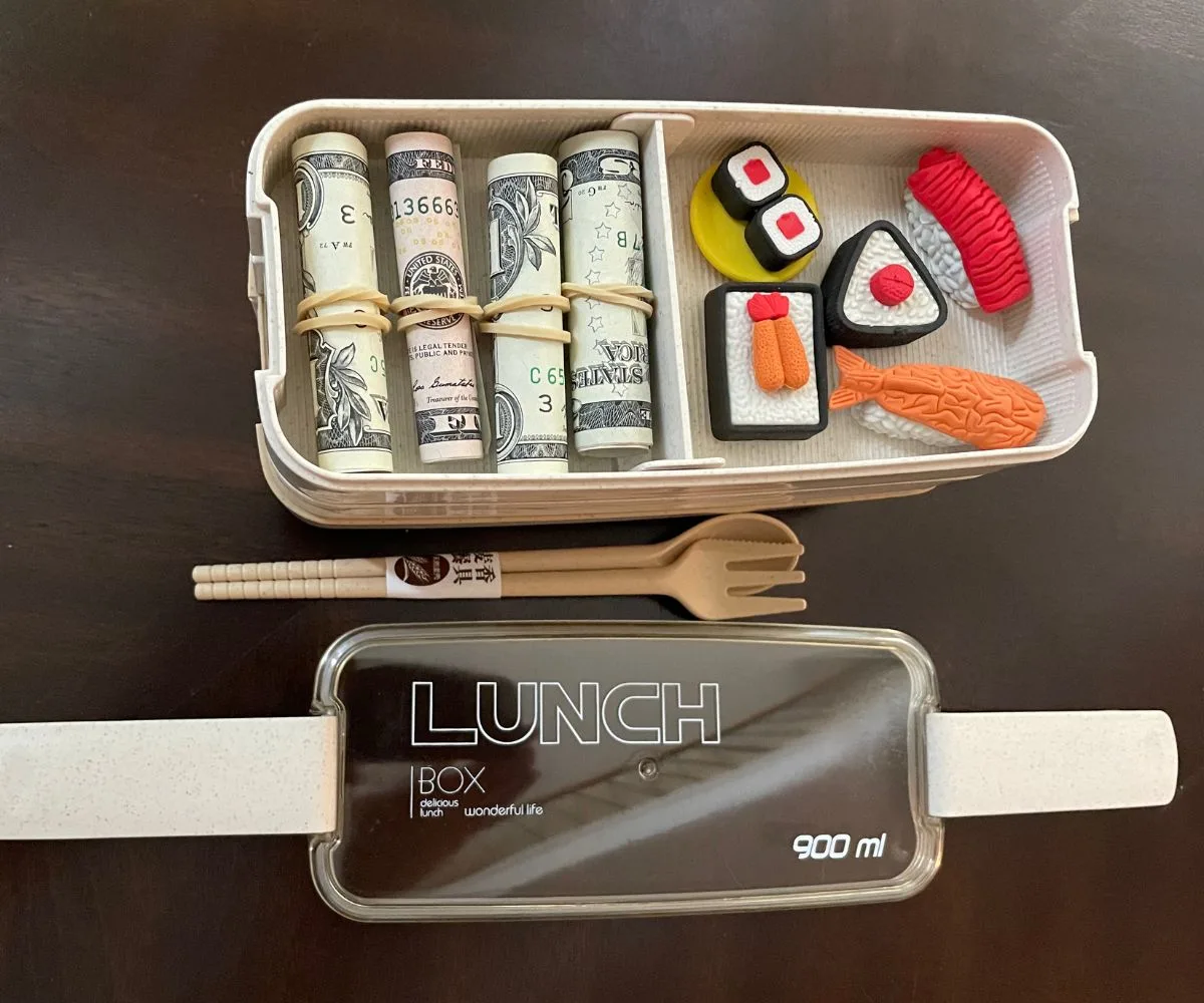three-tier bento box khaki colored with rolled dollar and 5 dollar bills on one part of top and eraser sushi in other, opened