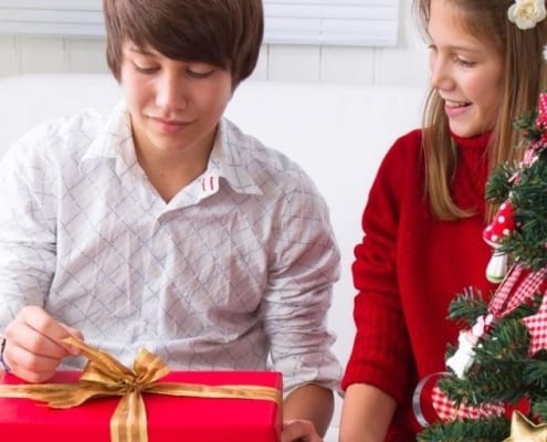 teenage guy smiling, opening up Christmas gift next to teen girl and Christmas tree