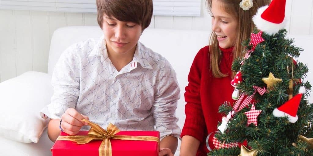 teenage guy smiling, opening up Christmas gift next to teen girl and Christmas tree