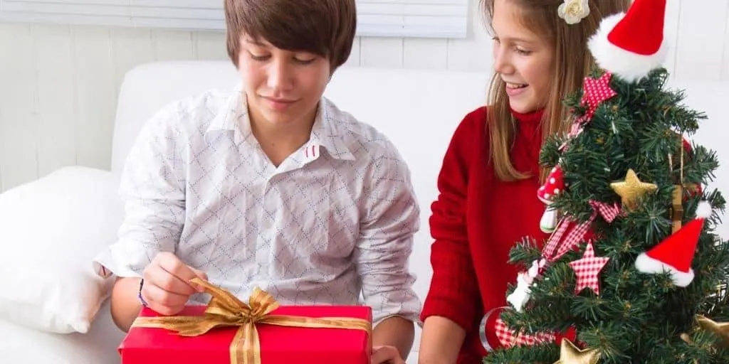 teenage guy smiling, opening up Christmas gift next to teen girl and Christmas tree