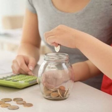 little girl putting coins into spend save share piggy bank money jar with mother and calculator