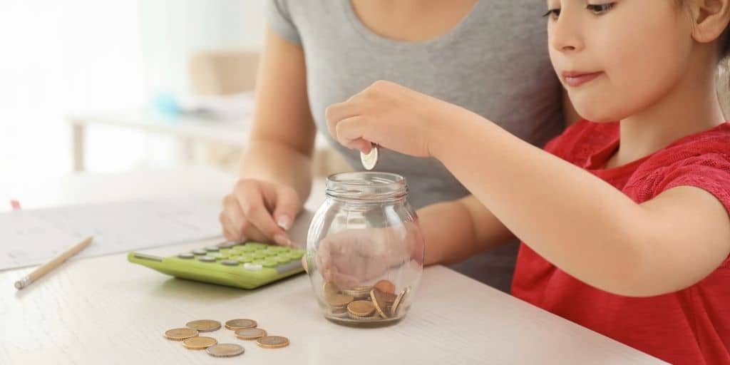 little girl putting coins into spend save share piggy bank money jar with mother and calculator