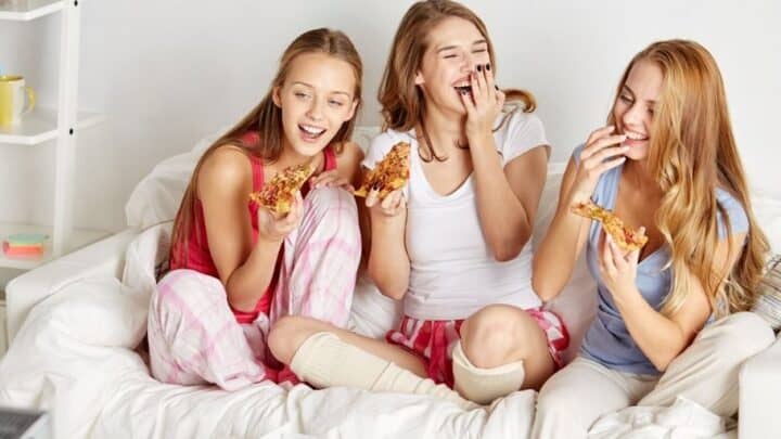 three teen girls laughing on bed with pizza doing 100 things to do at a sleepover at home