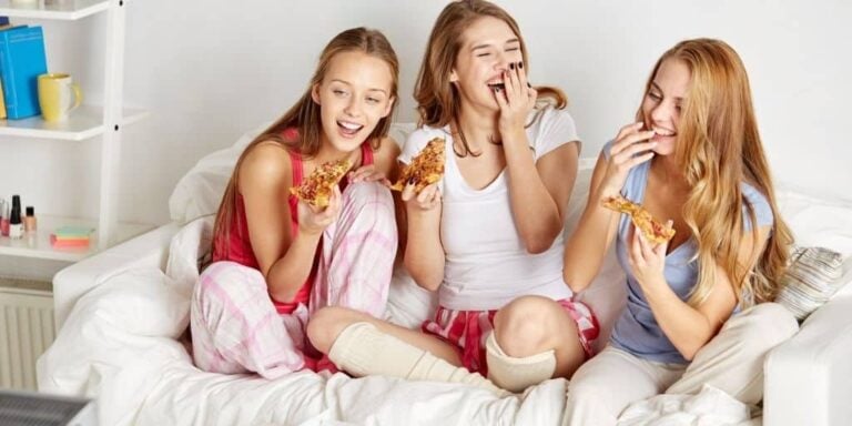 three teen girls laughing on bed with pizza doing 100 things to do at a sleepover at home