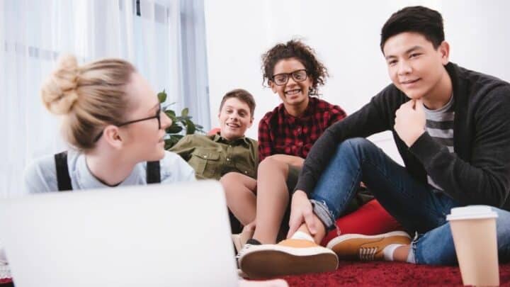 group of teens on ground hanging out, playing a stock market game for kids on laptop