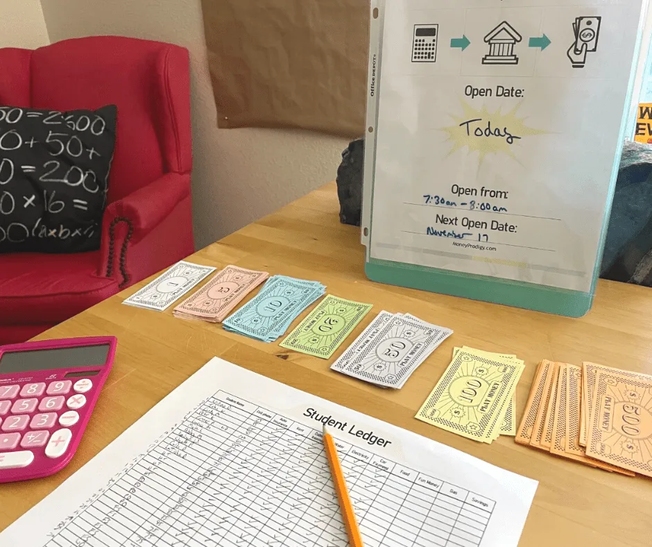 bill paying station on a table with a bill pay station sign that says open from 7:30 - 8:00 a.m. pretend money, calculator, and student ledger filled in