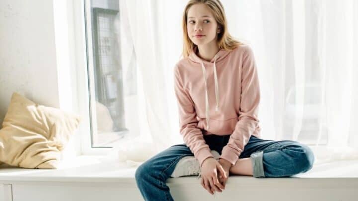 teen girl in pink sweatshirt on windowsill, looking annoyed