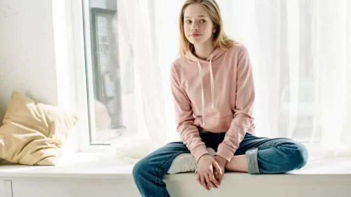 teen girl in pink sweatshirt on windowsill, looking annoyed