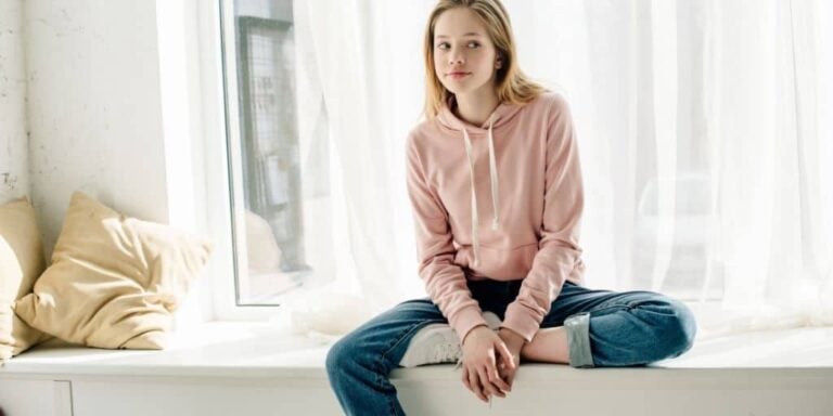 teen girl in pink sweatshirt on windowsill, looking annoyed
