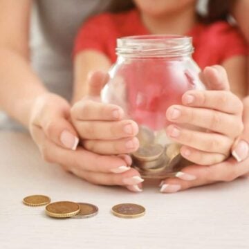 woman holding child's hand, who is holding a piggy bank with money