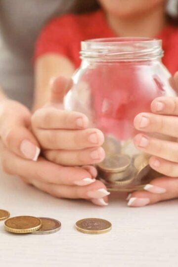 woman holding child's hand, who is holding a piggy bank with money