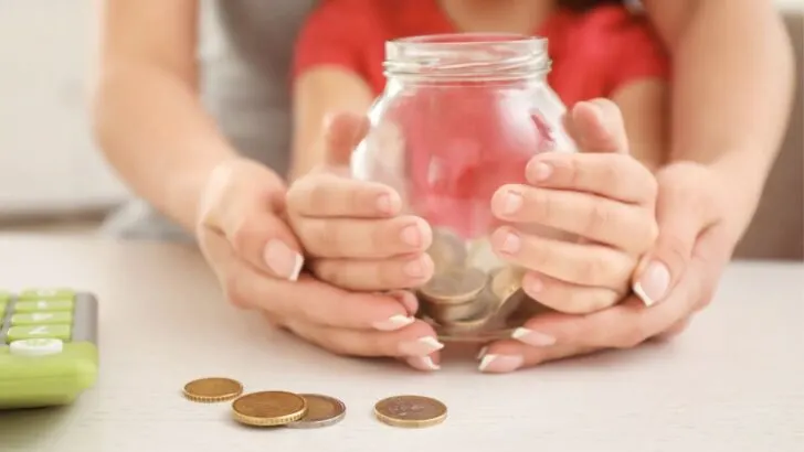woman holding child's hand, who is holding a piggy bank with money