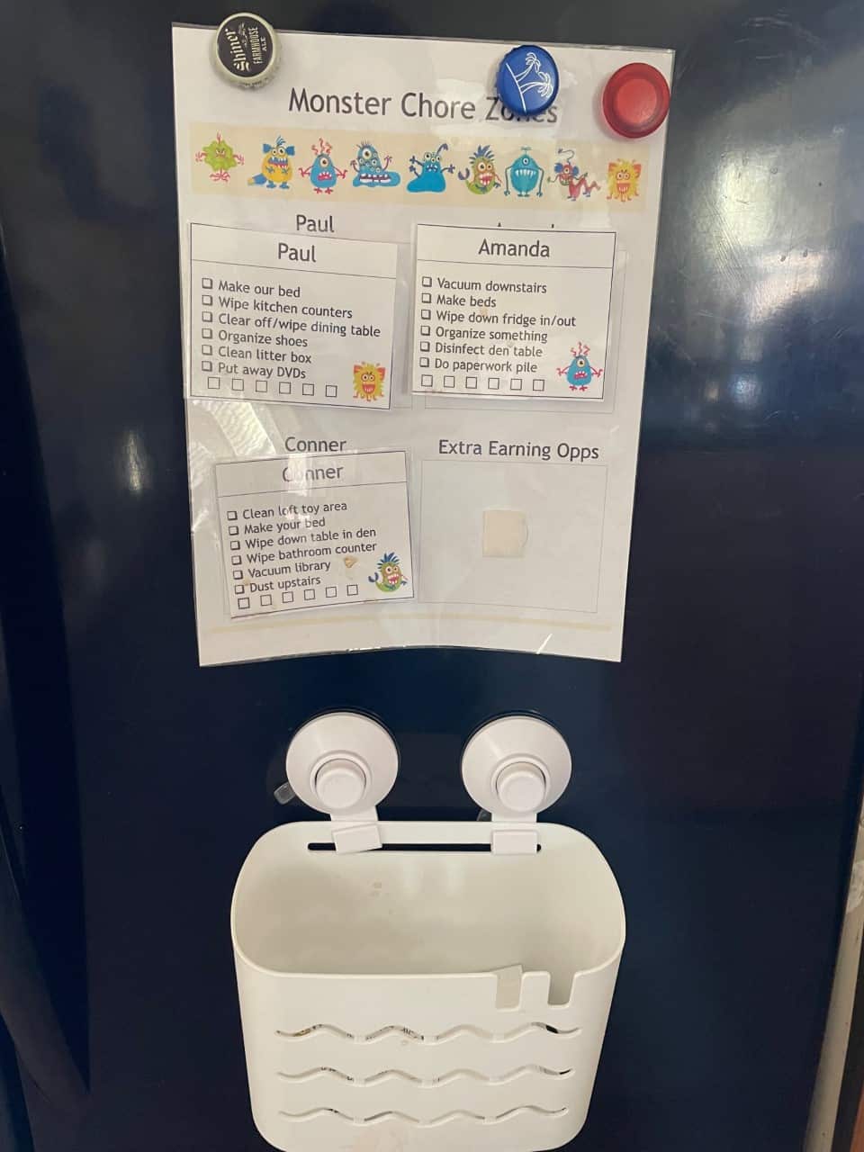 black fridge with monster chore chart with four squares on it for three people and extra earning opportunities, plus cards in a suction cup basket to rotate