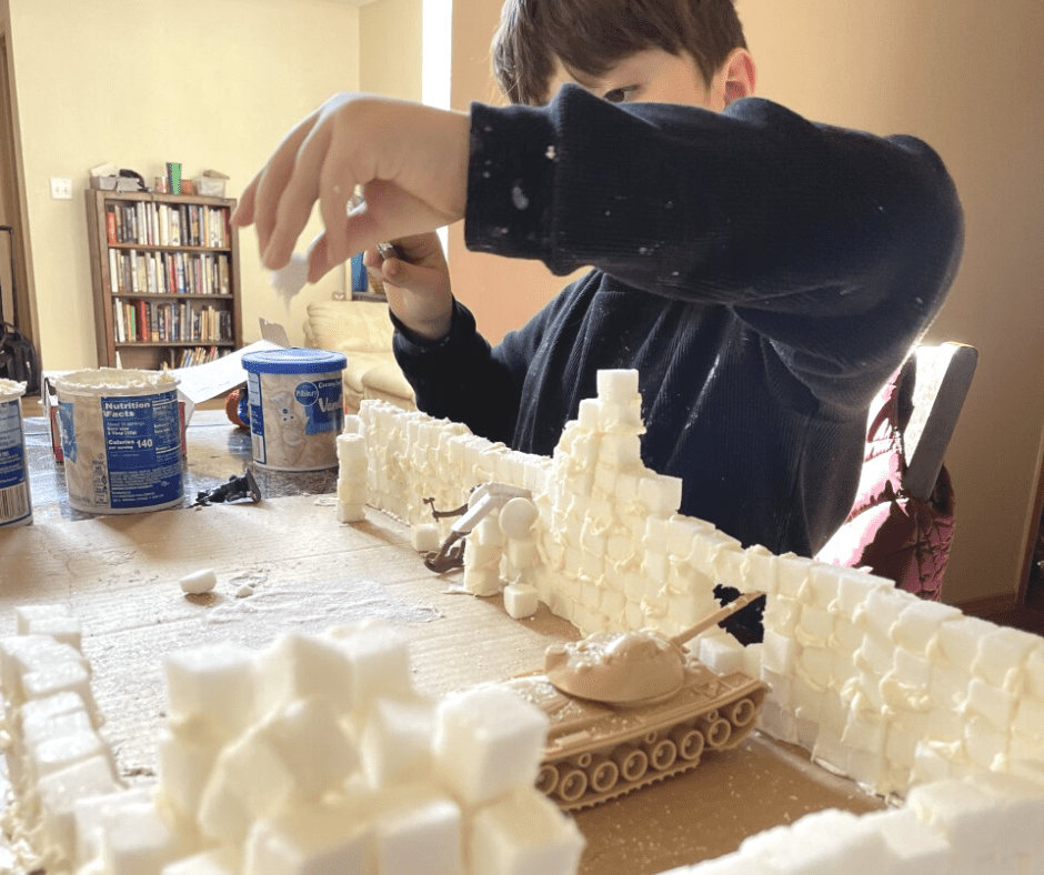 7-year-old boy in black shirt gluing sugar cube blocks with frosting on rectangular Alamo building he built
