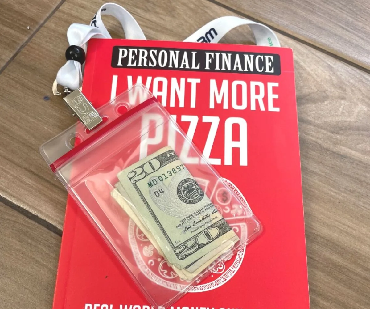 Red I Want Pizza book cover with employee lanyard, holding a $20 bill
