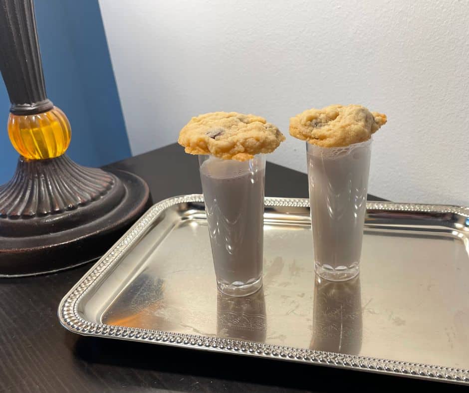 little shooter glasses of milk with a chocolate chip cookie on top, on silver tray