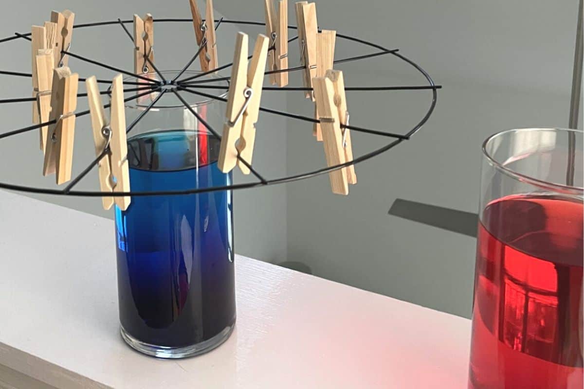 two glass vases filled with colored water, wire wreath frame sitting on top of one with clothespins hanging on it