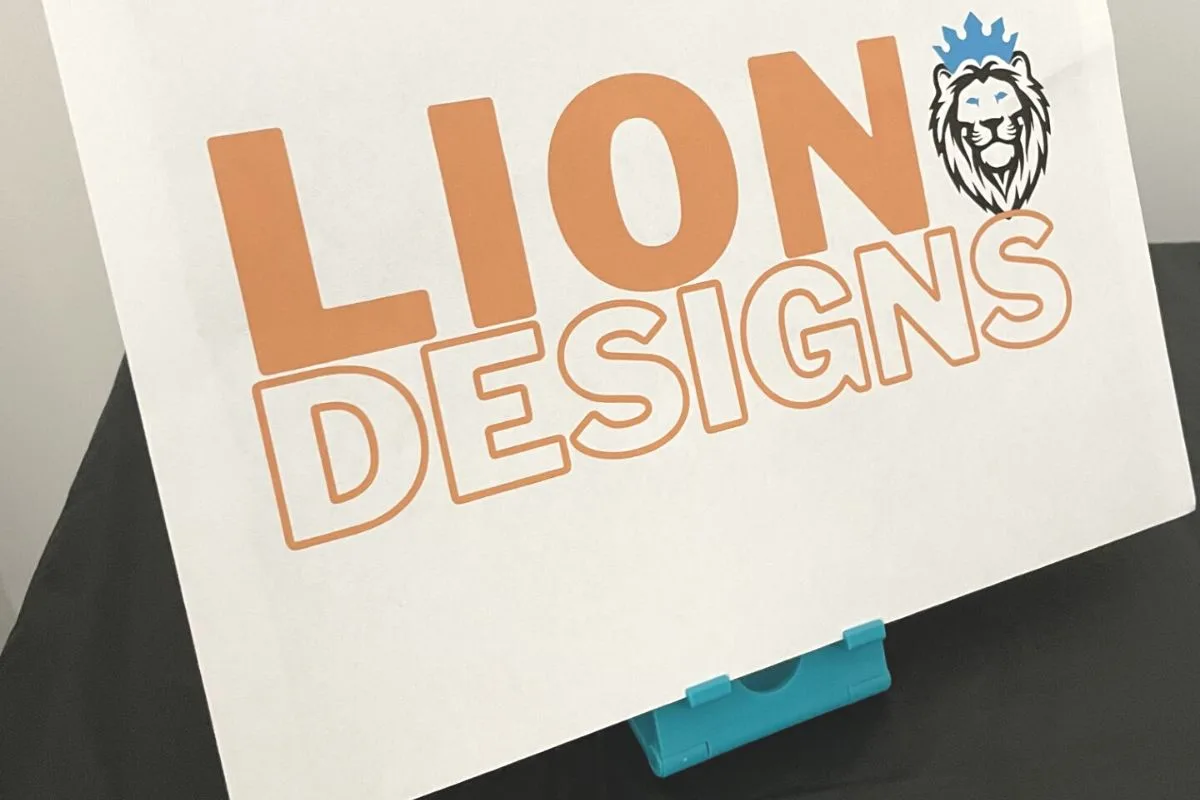 white paper propped up on blue phone stand, says "Lion Designs" with a lion and blue crown on its head