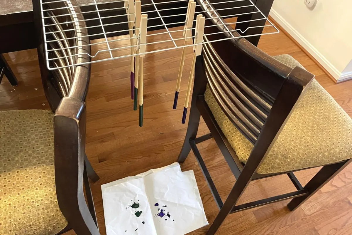 cooling rack bridging two chairs, with clothespins holding up chopsticks hanging over paper to catch dripped paint