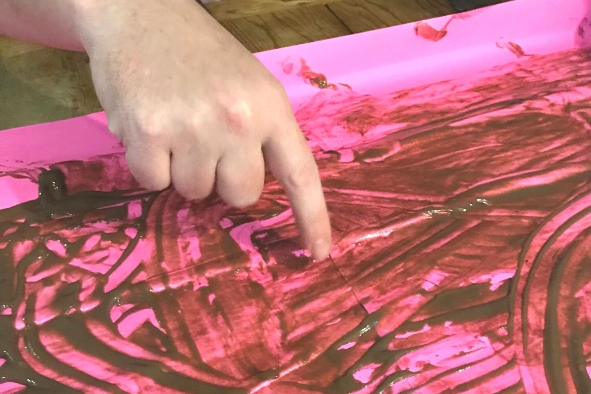 chocolate pudding on big fluorescent pink paper, with person's hand making a picture in it