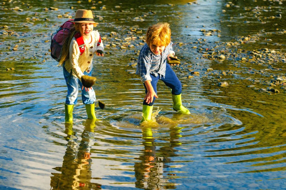 3 year old and 5 year old throwing big rocks into creek with rain boots on