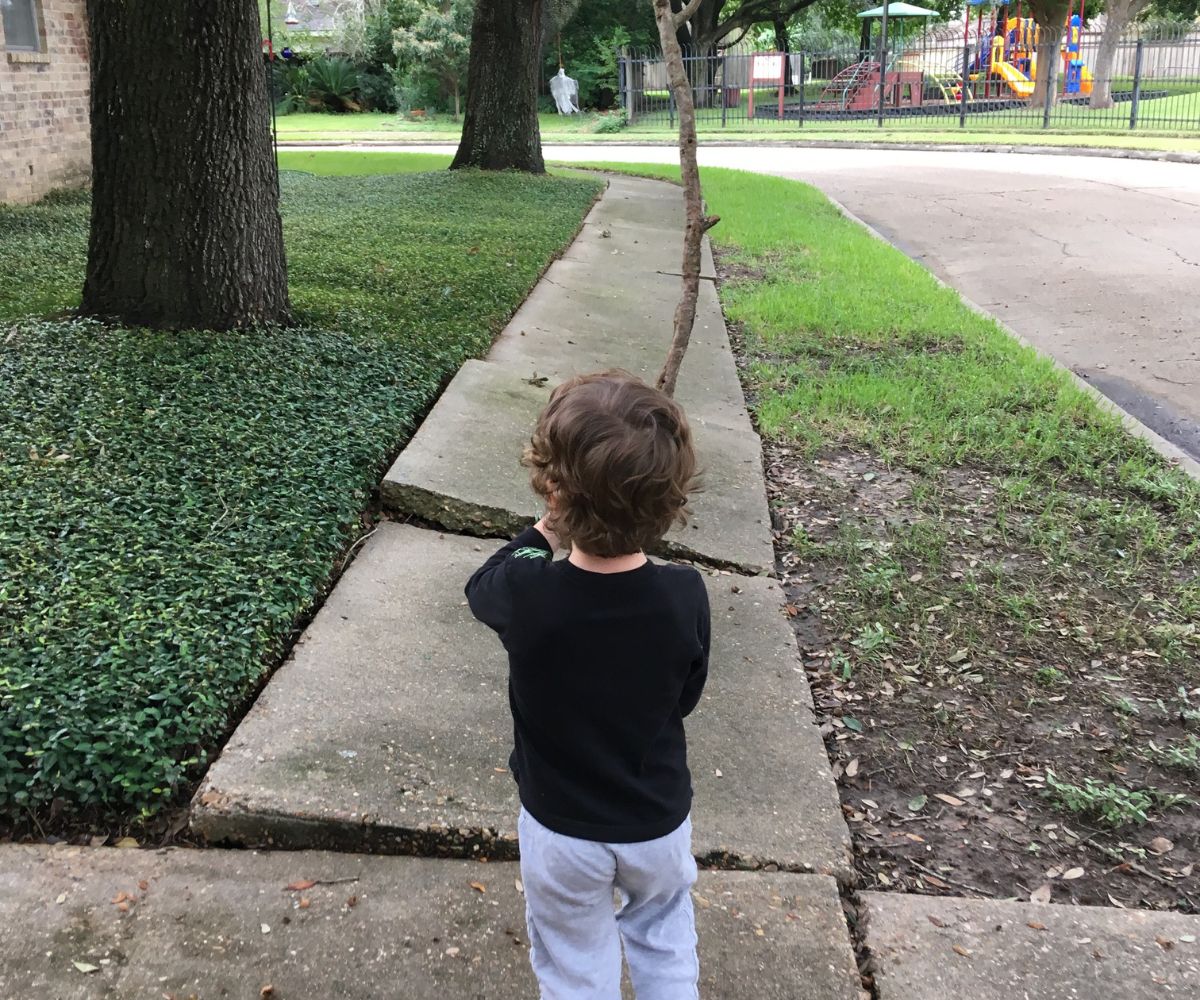 preschool boy outside with large stick in hand walking in front of mother to the park
