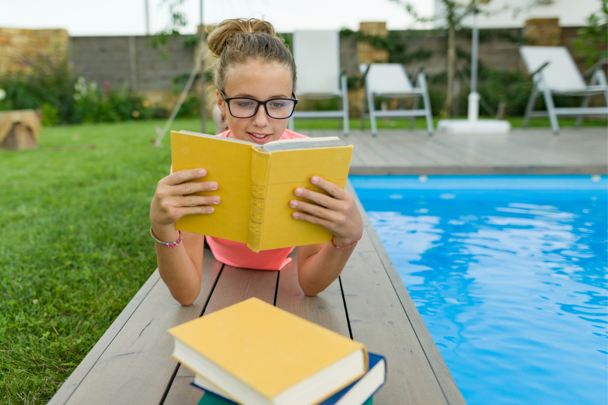 teen girl in glasses reading book alongside pool's edge
