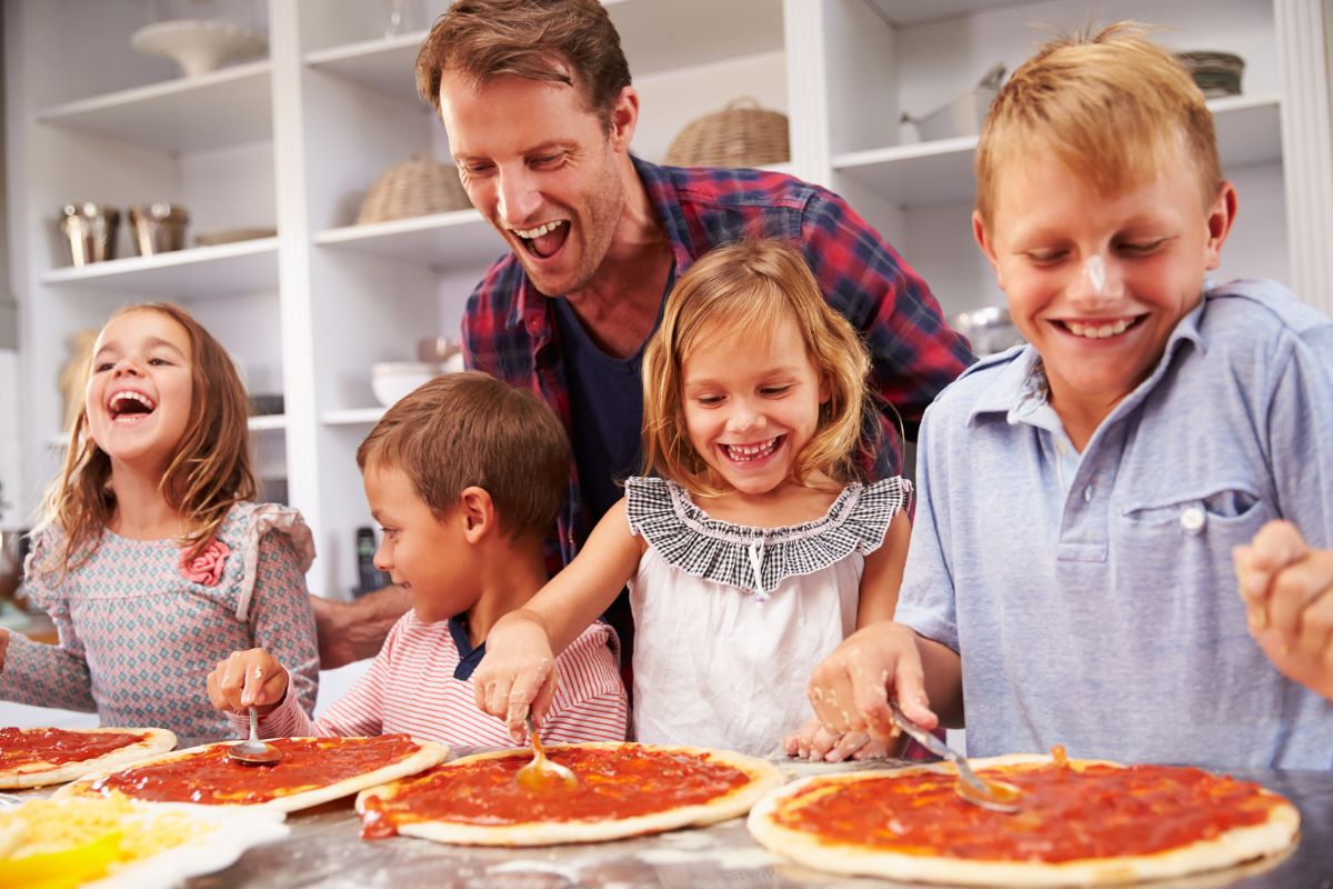 four kids putting sauce on four pizza doughs, father in back helping and laughing