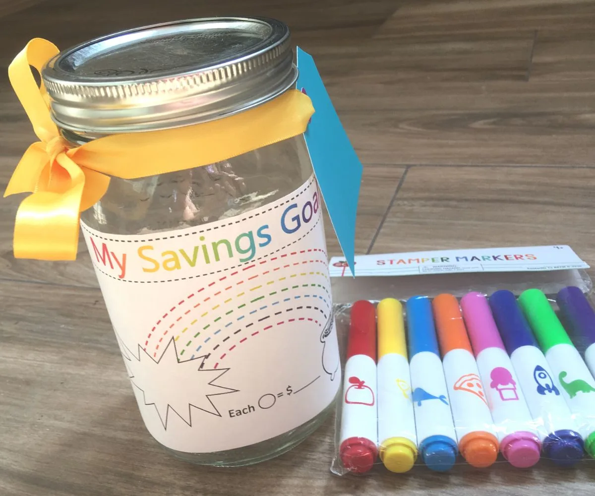 mason jar with My Savings Goal sticker on it, and yellow ribbon, next to stamp markers on ground
