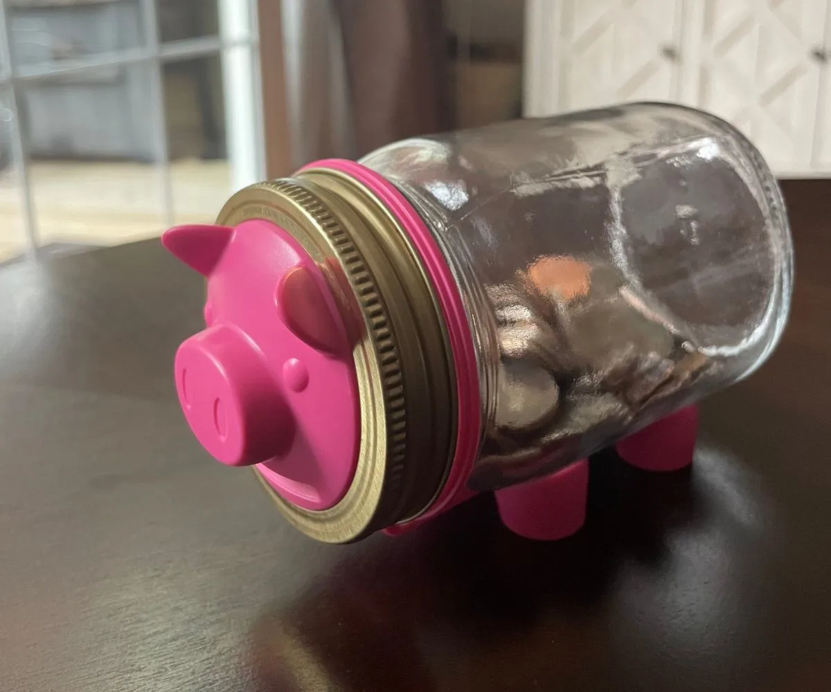 mason jar filled with coins on its side, with pink piggy face and pig feet holding it up
