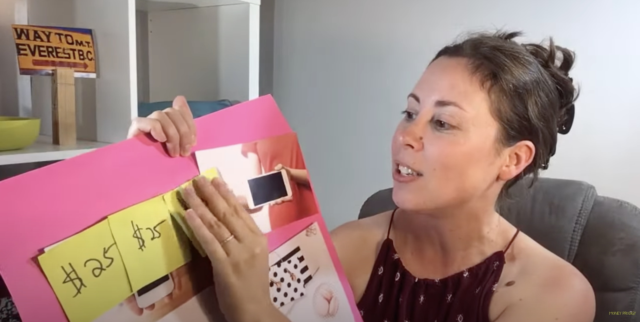 woman showing bright pink poster board with pictures of iphone on it, and $25 on three Post-It notes being placed on top of photos