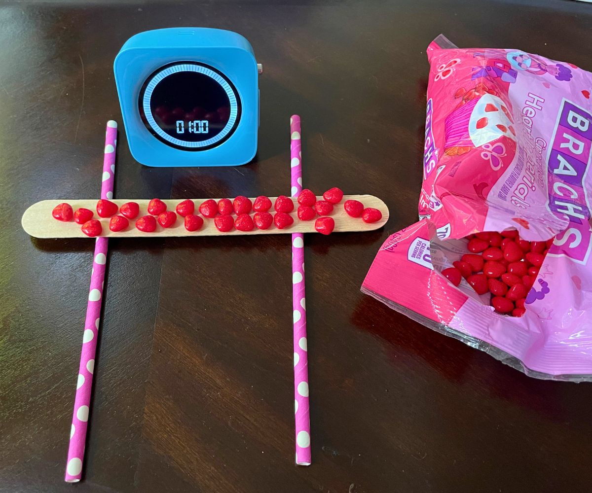 two pink polka dot straws with a large popsicle stick across them, filled with cinnamon heart candies, and a blue timer