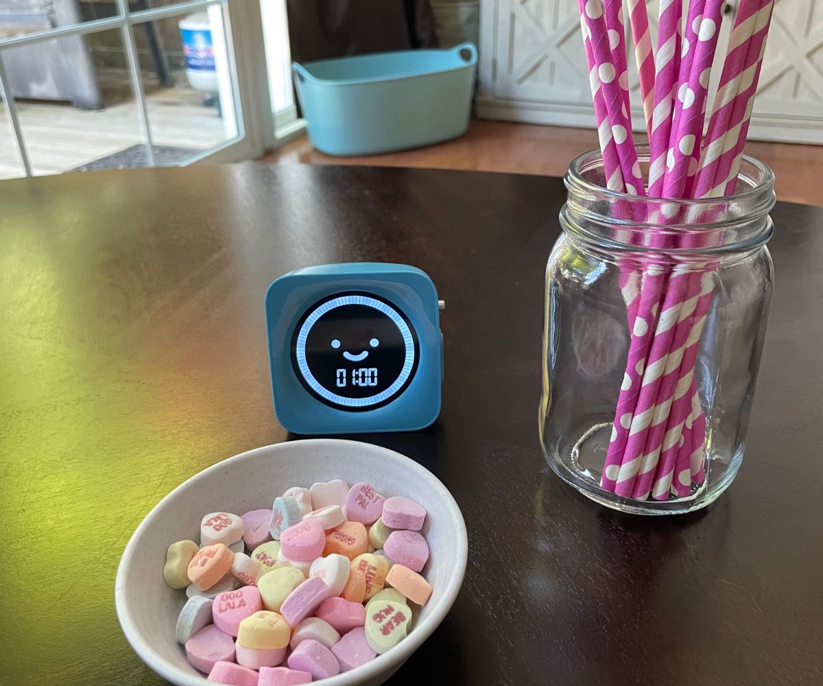mason jar of pink and white straws, blue timer, and little bowl full of candy conversation hearts