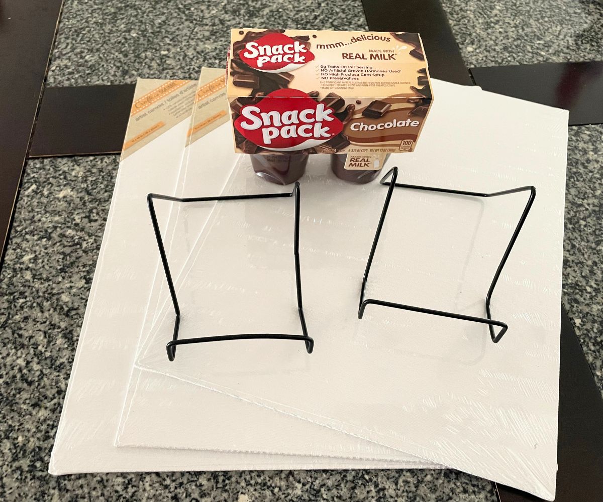 three canvases and canvas holders, plus a pack of Snack Pack puddings on top of table