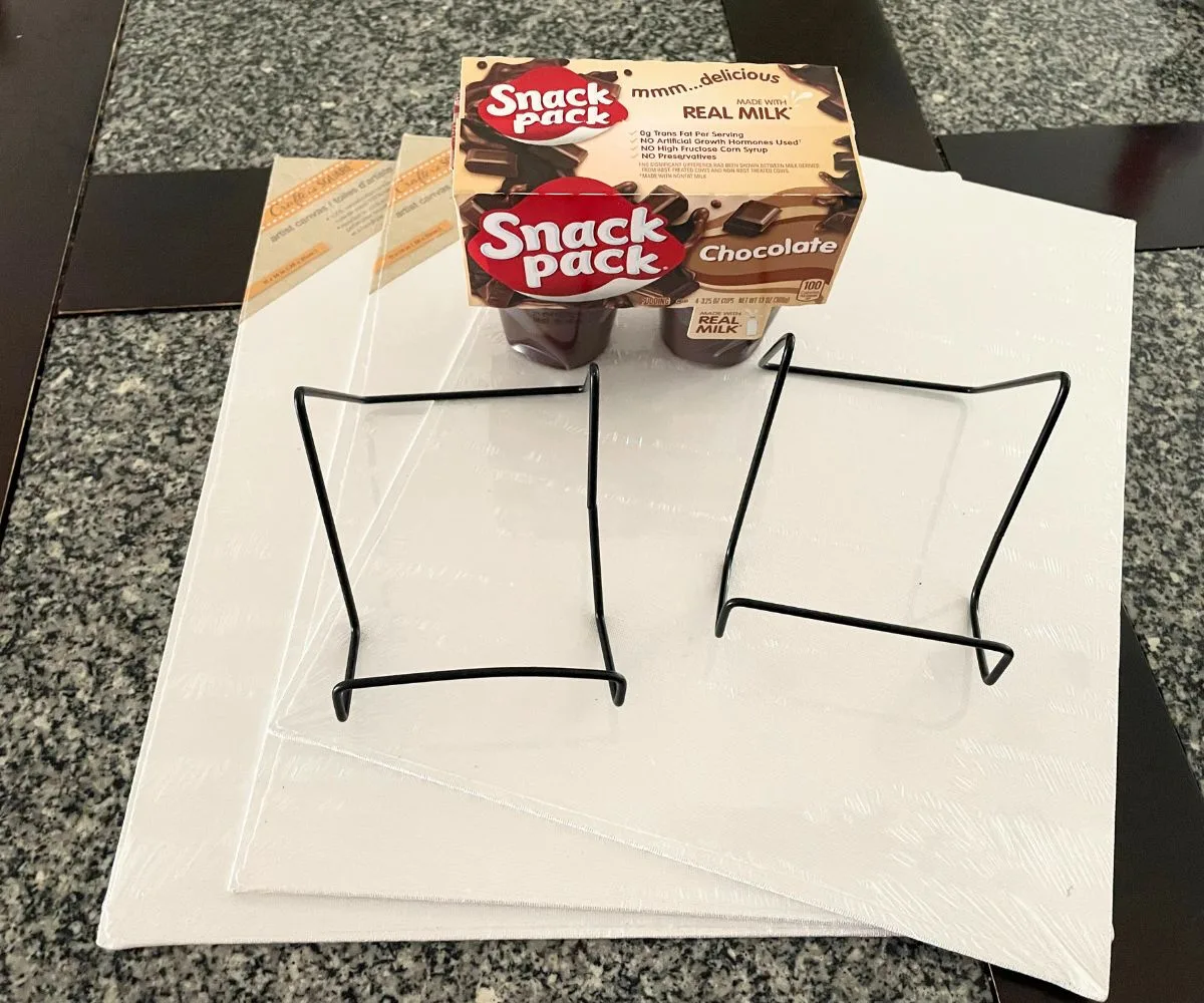 three canvases and canvas holders, plus a pack of Snack Pack puddings on top of table