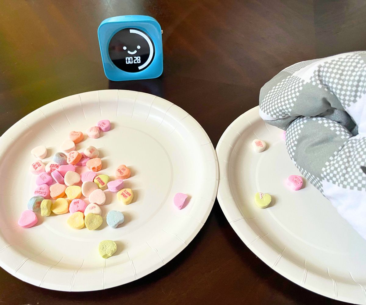 hand in oven mitt picking up one candy conversation heart at a time from one plate and moving to another, blue timer showing 28 seconds left