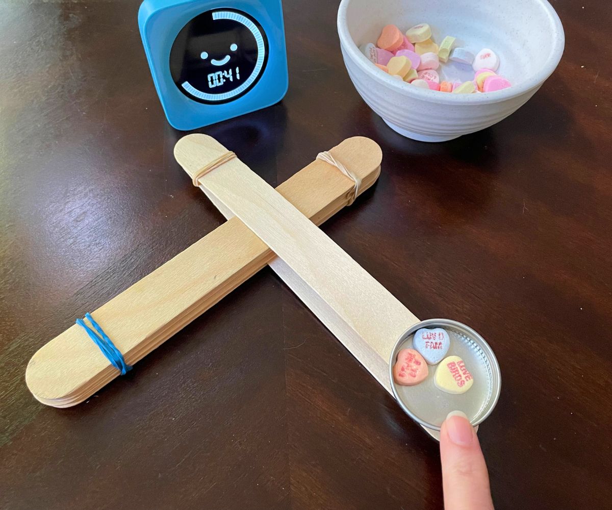 hoemmade catapult holding three candy conversation hearts with the words up, blue timer, on dark table