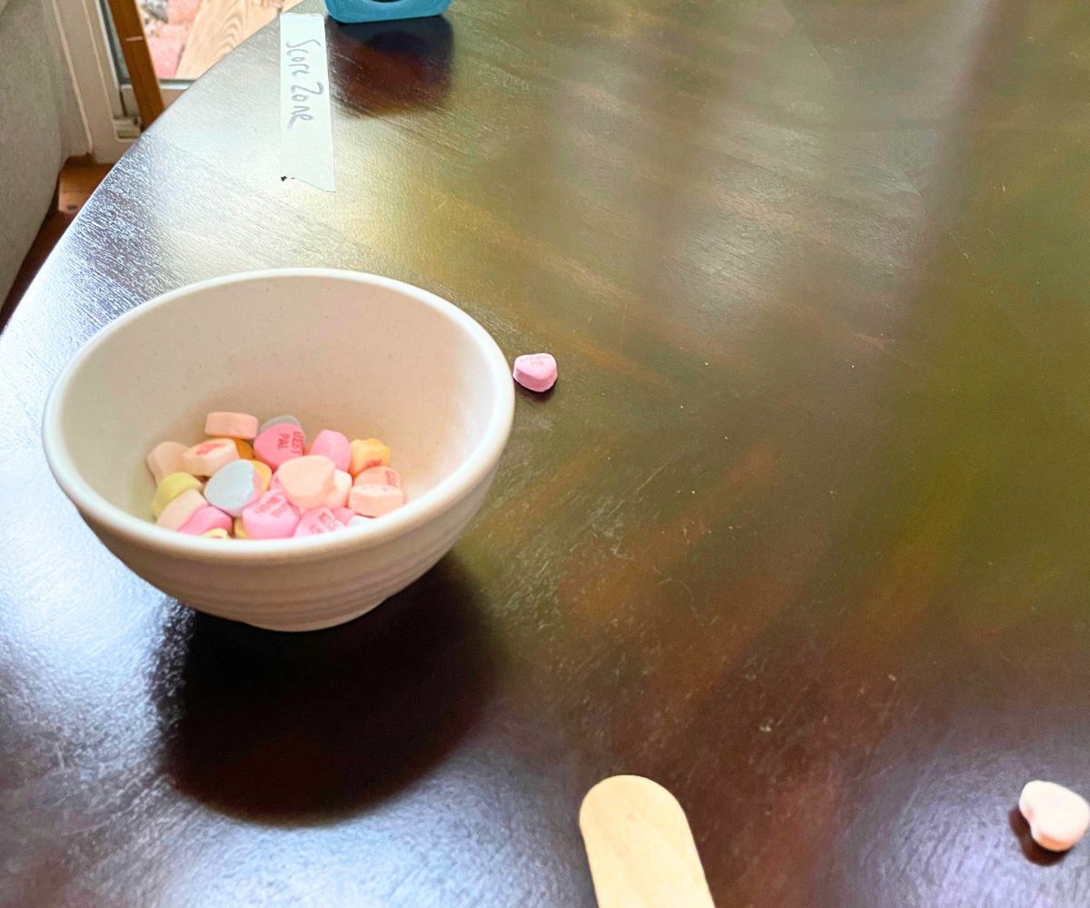 popsicle stick pushing candy heart across table, nowhere near the "Score Zone" labeled by a piece of masking tape