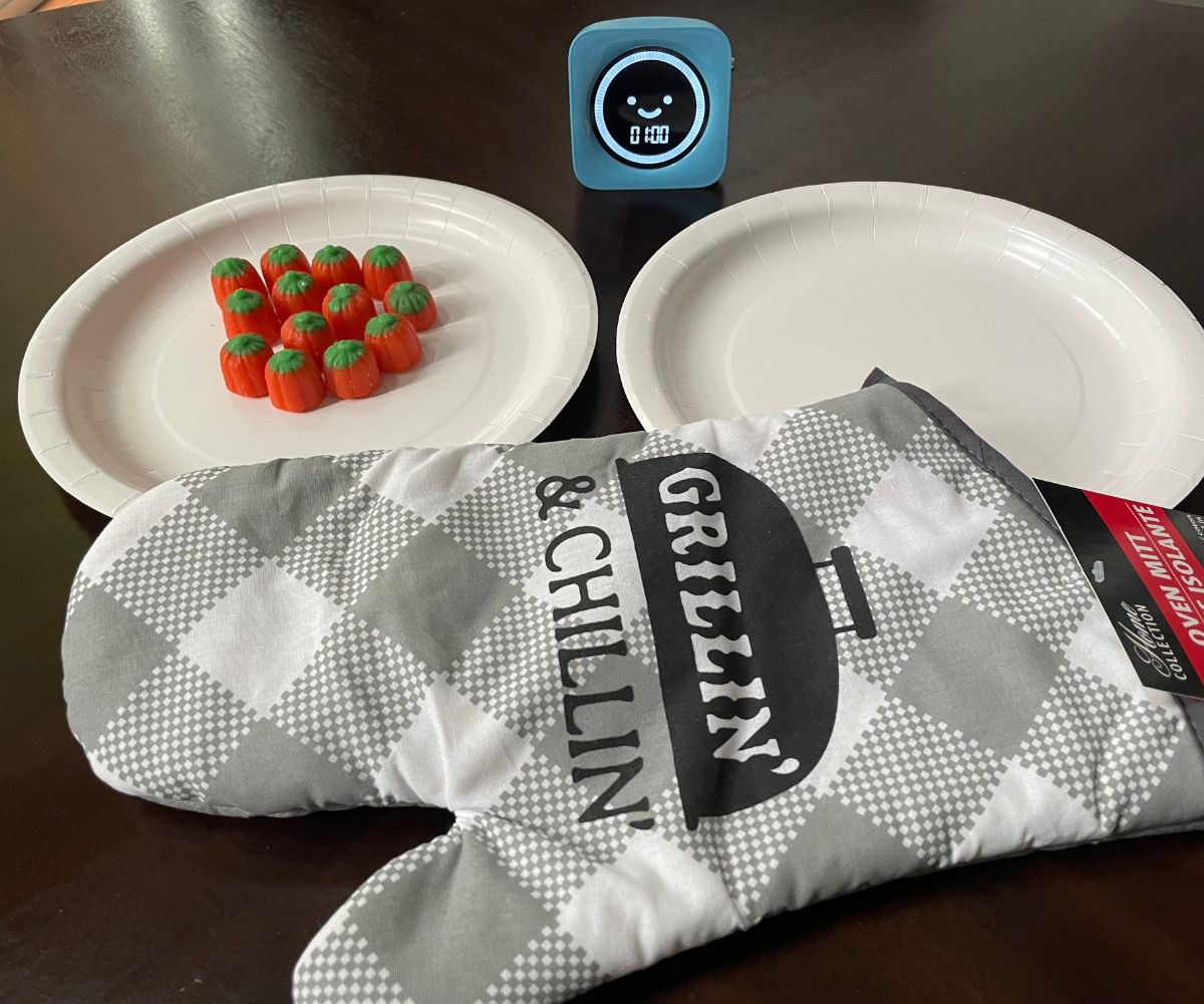 big oven mitt, two plates, one filled with candy pumpkins, and blue timer for 1 minute