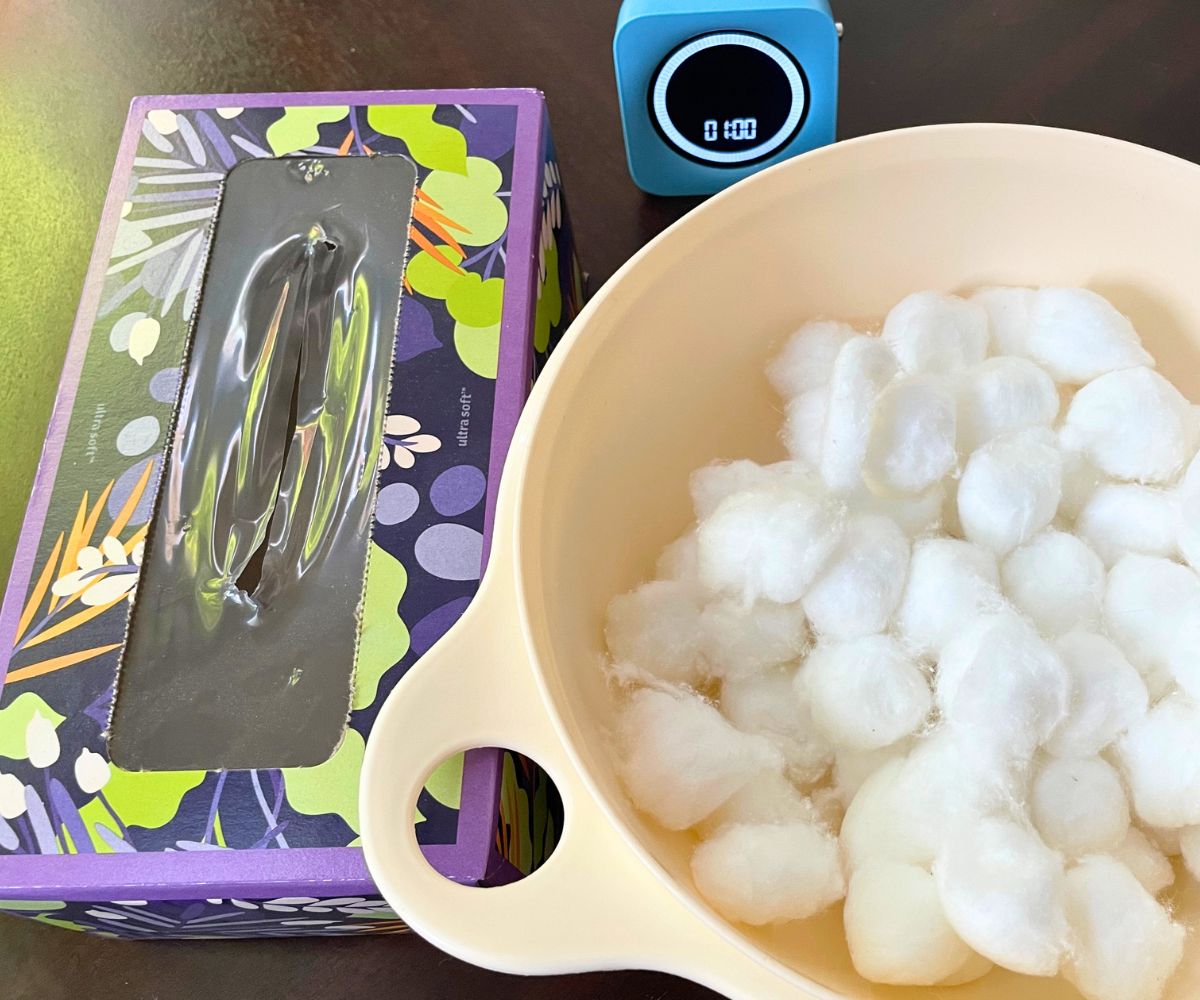 white bowl filled with cotton balls next to colorful empty tissue box and blue timer