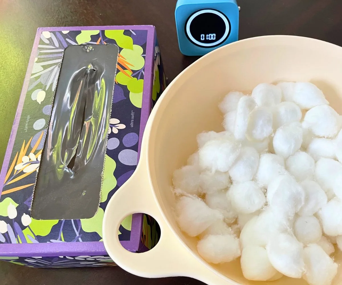 white bowl filled with cotton balls next to colorful empty tissue box and blue timer