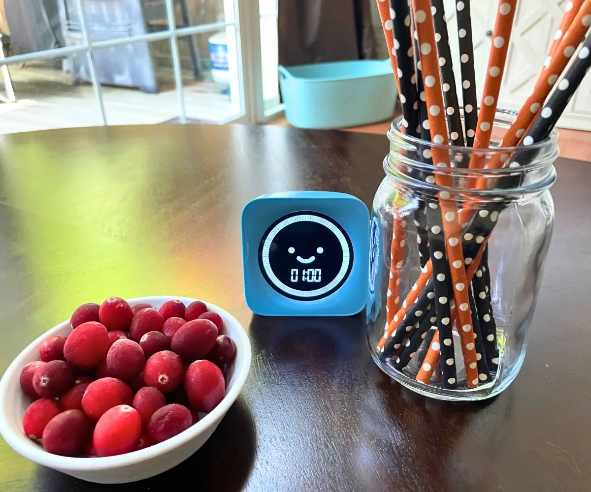 mason jar of orange and black straws, blue timer, and small bowl filled with bright red cranberries